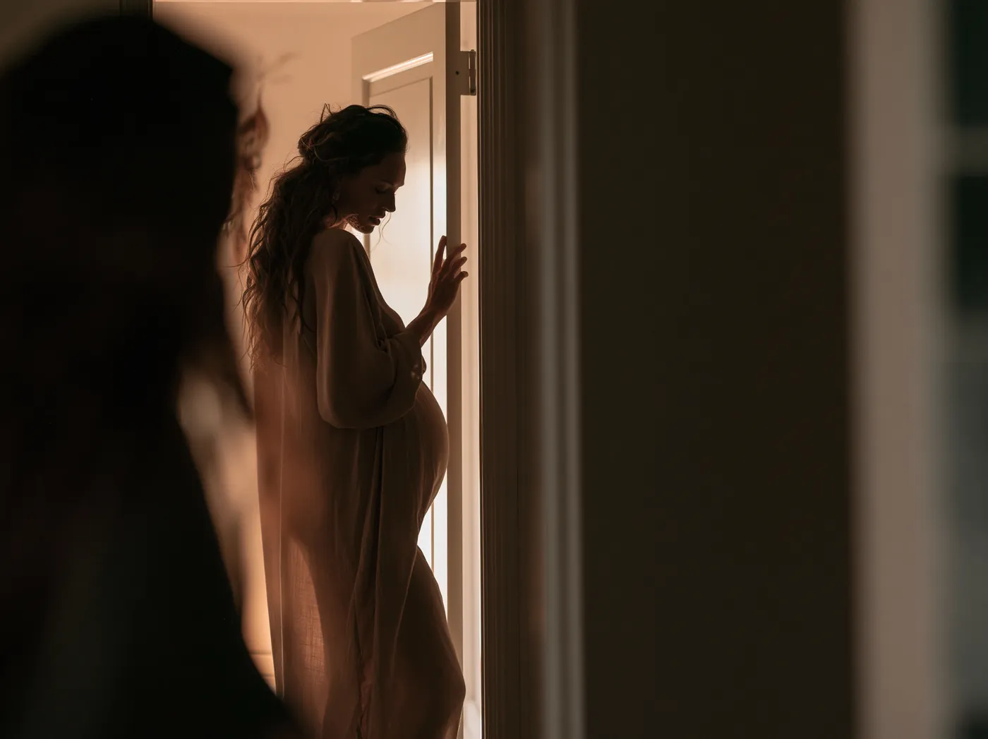 Silhouette of a pregnant woman resting a hand on a doorframe at home