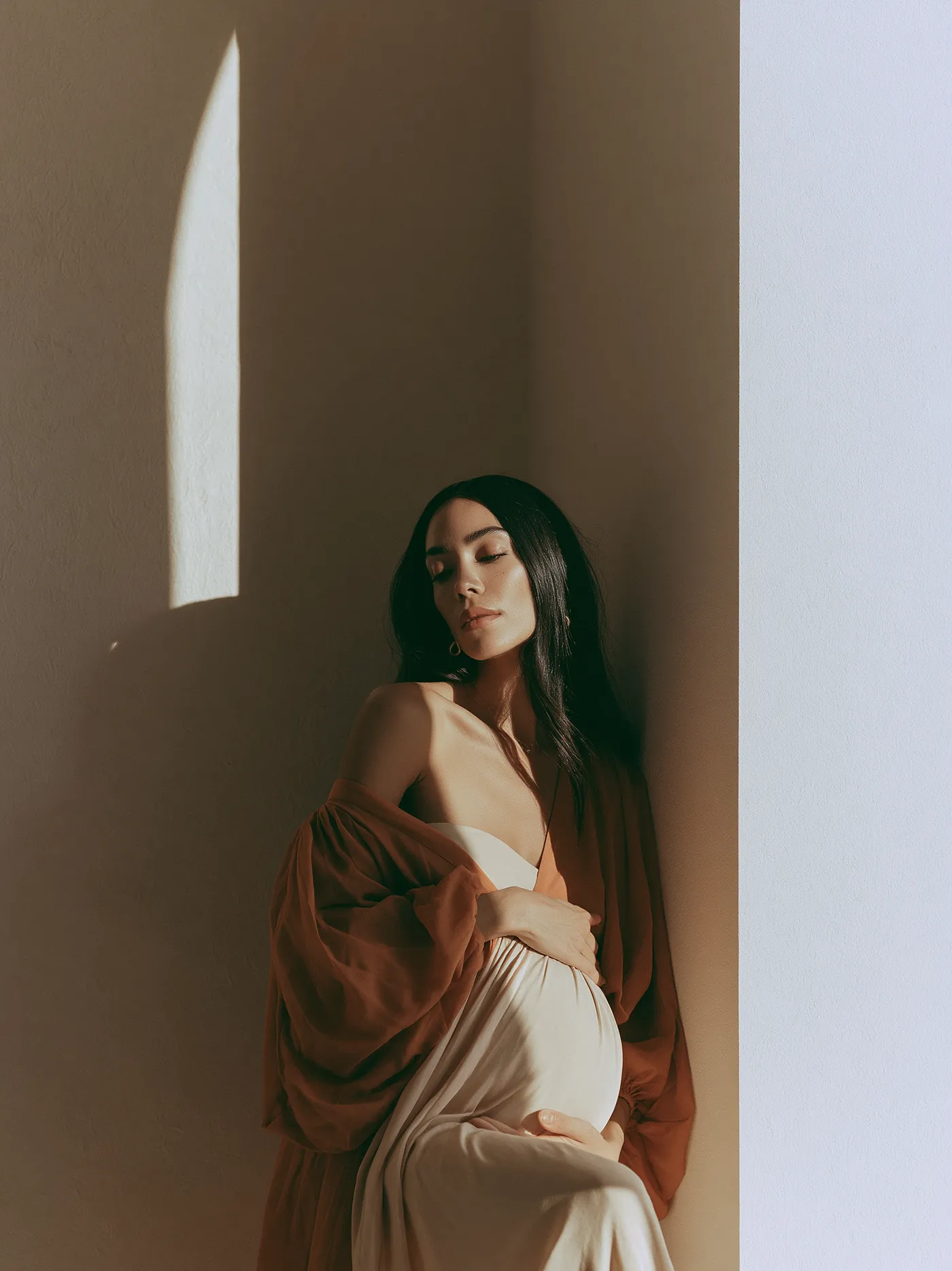 Woman in her third trimester leaning into a wall as afternoon light crosses it
